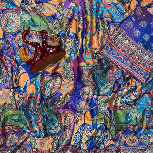 May include: A vibrant fabric featuring a multi-colored peacock and floral pattern in shades of blue, orange, and green. The textile includes a decorative border with geometric motifs and a pair of dark red, curved, swan-shaped ornaments.