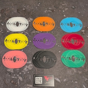 Op de afbeelding: Een verzameling kleurrijke, cirkelvormige speltoken in verschillende kleuren, waaronder wit, oranje, blauw, geel, paars, rood, zwart en groen. Elke token heeft een centraal gat en een gestileerd ontwerp. Een visitekaartje met een logo staat op de voorgrond.