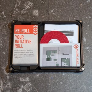 May include: A black, rectangular game organizer with a zipper closure. Inside are cards with text that reads "RE-ROLL YOUR INITIATIVE ROLL" and a map labeled "KILLZONE: VOLKUS". A red, circular game piece is also visible.