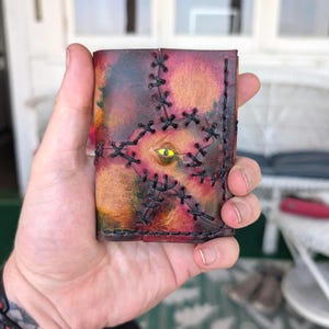 Puede incluir: Una cartera de cuero rectangular con una superficie multicolor moteada en tonos rojo, naranja y marrón oscuro. La cartera está acentuada con gruesas costuras cruzadas negras y un único ojo amarillo realista con una pupila negra.
