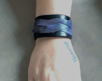 May include: A black leather cuff bracelet with a woven blue design. The bracelet is worn on a wrist. The word "Breathe" is tattooed on the wrist in blue ink.