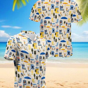 May include: Short-sleeved button-down shirt with a repeating pattern of beach scenes. The design includes ice cream stands, umbrellas, beach chairs, and trees in yellow, blue, and white on a cream background. The shirt is displayed on a beach setting.
