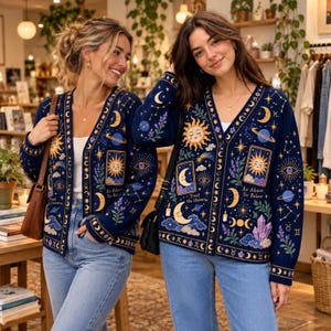 May include: A navy blue button-down cardigan featuring a celestial print with suns, moons, planets, stars, and crystals. The knit fabric includes the text "As Above, So Below" and "Trust the Universe" in a repeating pattern across the front.