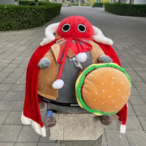 May include: A plush toy in the shape of a red-faced character with large black eyes, wearing a red cape with white trim, a brown jacket, and holding a burger-shaped pillow. The toy is sitting on a stone pedestal.