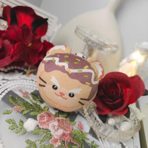 May include: A plush, round, tan-colored toy with a cartoon cat face and brown and green toppings. The toy has white ears and black whiskers. A red rose and other decorative elements are in the background.