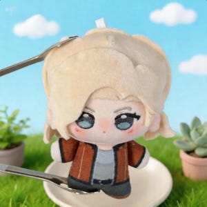 K&ouml;nnte beinhalten: Eine Pl&uuml;schpuppe mit hellblondem Haar und blauen Augen, die eine braune Jacke &uuml;ber einem grauen Hemd tr&auml;gt. Die Puppe befindet sich auf einem wei&szlig;en Teller, mit einem L&ouml;ffel und einer Gabel in der N&auml;he. Die Puppe hat ein niedliches Anime-Design.