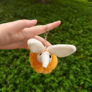 May include: A plush keychain featuring an orange, fuzzy body with white ears and a white trunk. The keychain has a silver metal ring attached to the top. The background is blurred green grass.