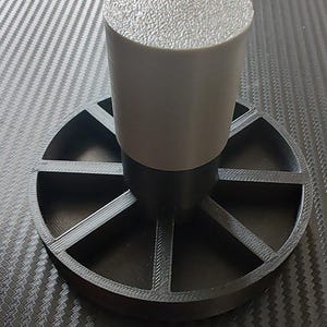 May include: A grey cylindrical component with a textured top, mounted on a black circular base with radial supports. The base rests on a dark, patterned surface. The object is likely a part of a larger assembly.