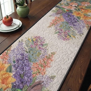 May include: A floral table runner with a cream background and a black border. The runner features a colorful design of flowers in shades of purple, pink, orange, and yellow. A hummingbird is also visible in the design. The runner is placed on a wooden table.