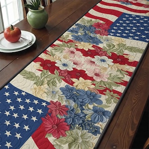 Puede incluir: Camino de mesa patriótico con diseño de la bandera estadounidense. Presenta rayas rojas y blancas, una zona azul con estrellas y motivos florales en rojo, blanco y azul. Colocado sobre una mesa de madera.