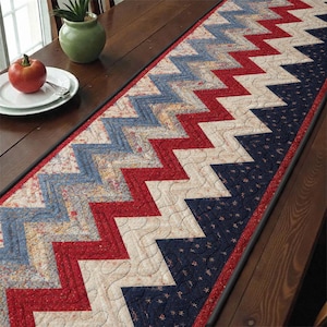 Puede incluir: Un camino de mesa acolchado con un patrón en zigzag en rojo, blanco y azul. El camino de mesa presenta un diseño patriótico con un borde rojo y está colocado sobre una mesa de madera. El camino de mesa mide aproximadamente 152 cm de largo.
