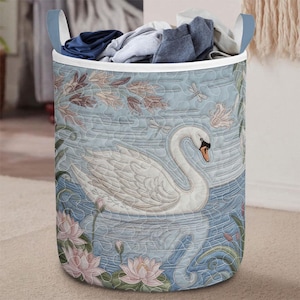Könnte beinhalten: Ein zylindrischer Wäschekorb mit hellblauem Hintergrund mit einem Schwanenmotiv. Der Korb hat einen weißen Rand, blaue Griffe und ist mit gefalteter Kleidung gefüllt. Der Schwan ist weiß mit einem orangefarbenen Schnabel, und das Design umfasst rosa Lotusblumen.