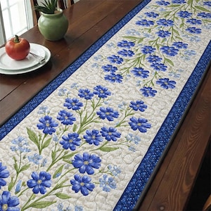 Puede incluir: Un camino de mesa floral con un fondo crema adornado con flores azules y hojas verdes. Tiene un borde azul y está colocado sobre una mesa de madera. El camino de mesa mide aproximadamente 152 cm de largo.
