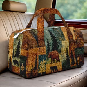 Op de afbeelding: Een gewatteerde duffeltas met een thema van de natuur. De tas heeft een patchworkpatroon met afbeeldingen van beren, dennenbomen en bladeren in bruin-, groen- en goudtinten. Hij heeft een witte rits en bruine handvatten.