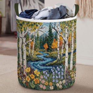 May include: A cylindrical laundry hamper with a forest scene design. The hamper features a quilted landscape with a river, trees, and flowers. The top edge is white, and the handles are dark green. The hamper is filled with folded clothes.