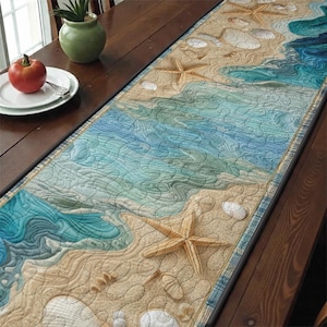 Może przedstawiać: Pikowany bieżnik na stół z motywem plaży. Bieżnik przedstawia piaszczystą plażę z muszlami i rozgwiazdami oraz fale oceanu w odcieniach niebieskiego i turkusowego. Bieżnik jest umieszczony na drewnianym stole.