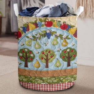 May include: A decorative laundry hamper with a quilted fruit and tree design. The hamper features a light blue background with apples, pears, and grape clusters. The bottom has a red and white checkered pattern.