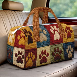 May include: A quilted duffel bag featuring a patchwork design with paw prints in various colors. The bag has a white zipper and brown handles. The color palette includes red, yellow, blue, and cream. The bag is sitting on a car seat.