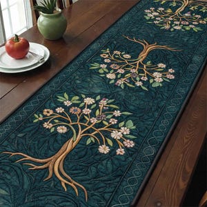 May include: A teal table runner with a tree of life design. The runner features embroidered trees with tan trunks, green leaves, and white and pink flowers. The runner is on a dark wooden table.