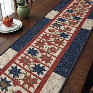 May include: A quilted table runner with a patriotic theme. The runner features alternating red and blue star patterns, floral accents, and a navy blue dotted border. The runner is rectangular and appears to be made of fabric.