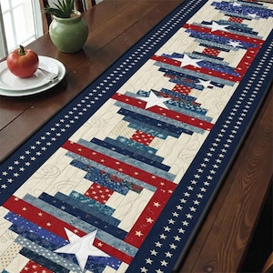 May include: A patriotic table runner featuring a patchwork design with red, white, and blue fabrics. The runner has a central pattern of log cabin blocks and large white stars, framed by a navy border with white stars. The runner is approximately 60 inches long.