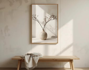 May include: A framed print featuring a beige vase with dried branches. The artwork is displayed on a light-colored wall above a wooden bench with a beige throw blanket. The scene is bathed in natural light, creating soft shadows.