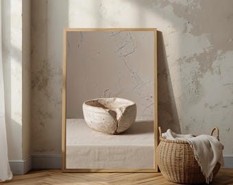 May include: A framed print featuring a textured, off-white ceramic bowl with a unique, cracked design. The bowl sits on a neutral-colored surface, with a woven basket and blanket in the background. The frame is a light wood color.