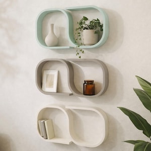 May include: Three modern, wall-mounted shelves in a light colour palette. The top shelf is a soft green and holds a white vase and a potted plant. The middle shelf is grey with a framed photo and a brown jar. The bottom shelf is white with books.