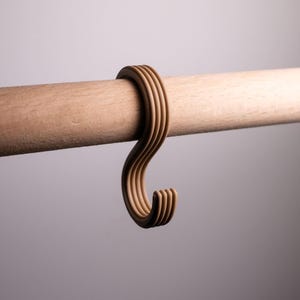 May include: A light brown wooden hook, with a curved design, hangs from a wooden rod. The hook has a series of curved lines. The rod is also light brown. The background is a soft grey.