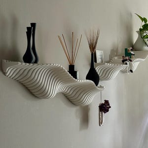 May include: White, wave-shaped wall shelves with black vases and decorative items. The shelves have a layered design, and the items include dried plants and a small framed picture. The shelves are mounted on a light gray wall.