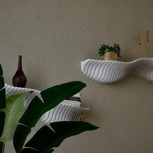 May include: A modern, wavy white shelf with a textured surface is mounted on a neutral-colored wall. The shelf holds small potted plants, gold candlesticks, and a decorative vase. A large green plant with broad leaves is in the foreground.