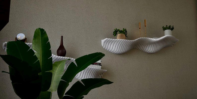 May include: A modern, wavy white shelf with a textured surface is mounted on a neutral-colored wall. The shelf holds small potted plants, gold candlesticks, and a decorative vase. A large green plant with broad leaves is in the foreground.
