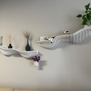 May include: Two white, wave-shaped wall shelves with black vases, dried plants, and a small potted plant. The shelves are mounted on a light gray wall, creating a modern, minimalist aesthetic. The shelves are ideal for displaying decorative items.