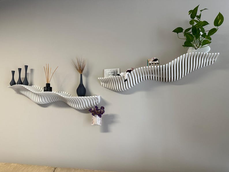 May include: Two white, wave-shaped wall shelves with black vases, dried plants, and a small potted plant. The shelves are mounted on a light gray wall, creating a modern, minimalist aesthetic. The shelves are ideal for displaying decorative items.
