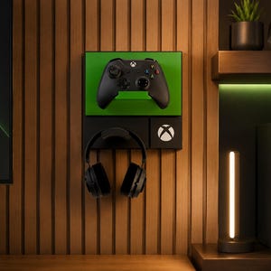 May include: A black wall-mounted gaming organiser featuring a green rectangular panel for a black wireless controller, a dedicated hook for black over-ear headphones, and a small square panel displaying the white circular Xbox logo emblem.