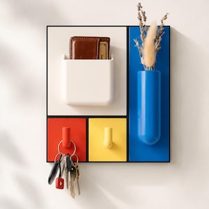 May include: A contemporary wall organiser with a black frame. It features a white pocket holding a brown wallet, a blue vase with dried flowers, and red and yellow hooks. Keys hang from the red hook. The design is geometric and colourful.