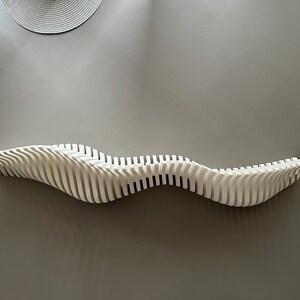 May include: A white, wave-shaped wall shelf made of vertical slats. The shelf is mounted on a gray wall, with a circular woven wall hanging above it. The shelf's design is modern and minimalist.