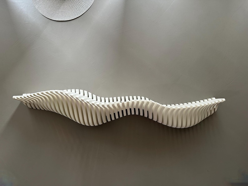 May include: A white, wave-shaped wall shelf made of vertical slats. The shelf is mounted on a gray wall, with a circular woven wall hanging above it. The shelf's design is modern and minimalist.