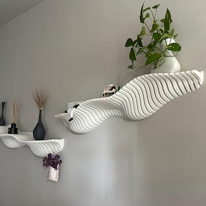 May include: Three white, wave-shaped wall shelves are mounted on a gray wall. The shelves hold decorative items, including black vases, dried plants, and a potted green plant. The shelves have a unique, modern design.