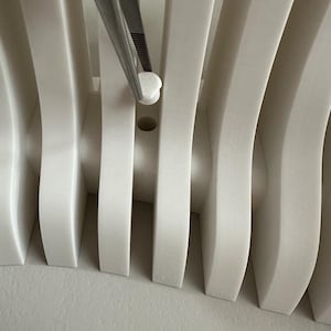 May include: Close-up of a white, curved architectural element with vertical supports. A small, white, rounded object is held by tweezers, positioned near a hole in the structure. The design features a repeating pattern of curved lines.