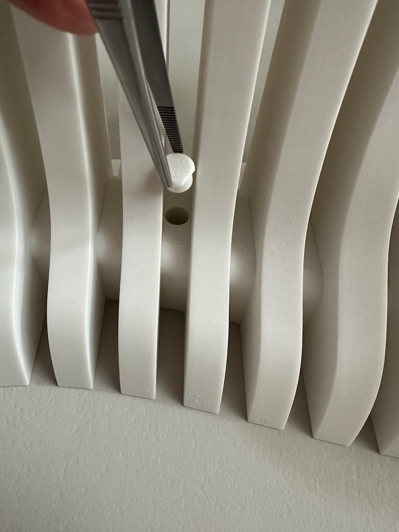 May include: Close-up of a white, curved architectural element with vertical supports. A small, white, rounded object is held by tweezers, positioned near a hole in the structure. The design features a repeating pattern of curved lines.