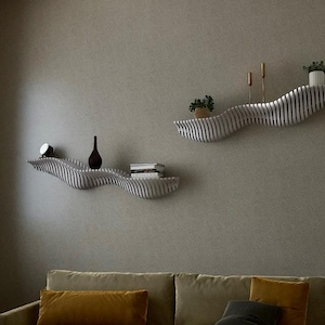 May include: Two modern, wavy wall shelves in a living room setting. The shelves are light-colored with a striped design, holding decorative items like a small clock, a dark vase, books, and potted plants. A sofa and a large window are also visible.