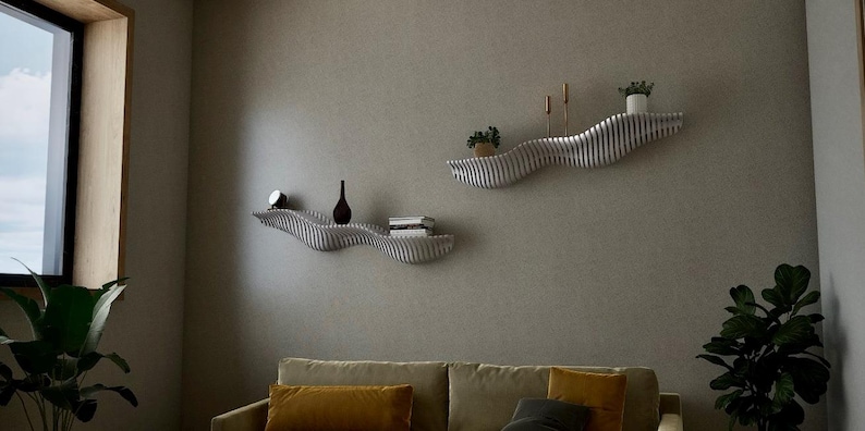 May include: Two modern, wavy wall shelves in a living room setting. The shelves are light-colored with a striped design, holding decorative items like a small clock, a dark vase, books, and potted plants. A sofa and a large window are also visible.