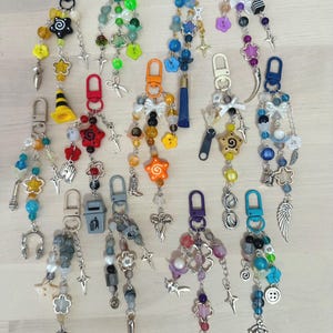 May include: A collection of thirteen colourful keychains featuring various plastic and metal charms, including stars, flowers, bows, and miniature objects like a traffic cone, trash can, hammer, and theatre masks, all attached to plastic lobster clasps.