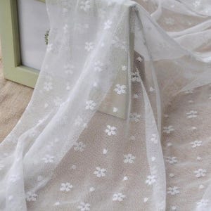 May include: White sheer fabric featuring a delicate floral pattern. The fabric is draped, showcasing its texture and the intricate design of small, evenly spaced flowers. The material appears lightweight and airy, suitable for various craft projects.