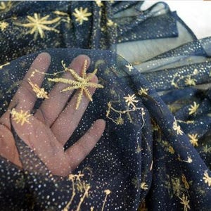 Könnte beinhalten: Dunkelblauer Stoff mit goldener Stern- und Sternbildstickerei. Das transparente Material ist mit kleinen goldenen Punkten übersät, die einen himmlischen Effekt erzeugen. Der Stoff ist gerafft und zeigt seine fließende Textur.