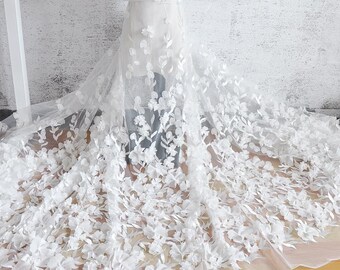 May include: White bridal fabric with 3D floral embellishments. The fabric is sheer, with a delicate, flowing drape. The flowers are clustered across the fabric, creating a textured, romantic look. The fabric is likely used for wedding dresses or other formal wear.