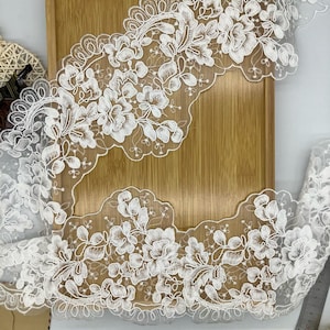 May include: White floral lace trim with a scalloped edge. The delicate lace features detailed embroidered flowers and leaves. The trim is ideal for sewing, crafting, and adding a decorative touch to garments or home decor projects.