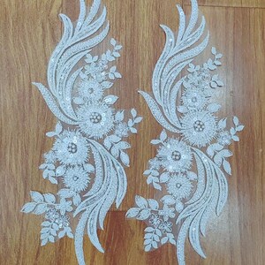 May include: Two white lace appliques with floral and leaf designs. Each applique features a central flower surrounded by smaller blossoms, leaves, and delicate sequin embellishments. The appliques have a symmetrical, flowing shape.