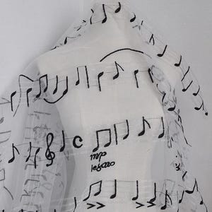 Op de afbeelding: Transparante witte stof met een patroon van zwarte muzieknoten. De stof is gedrapeerd en toont het delicate ontwerp. De tekst "mp legato" is op de stof gedrukt. Deze stof is geschikt voor het maken van unieke kleding of decoratieve items.
